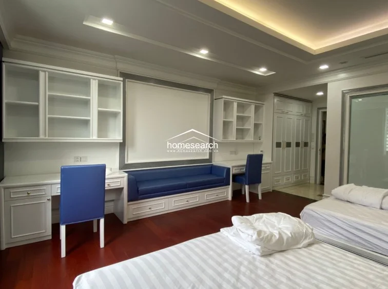This versatile room functions as both a bedroom and a study, featuring twin beds and a dedicated workspace with built-in desks, shelving, and a blue upholstered bench. The space is finished with rich, dark wood flooring and white cabinetry, providing a clean, organized, and functional layout.