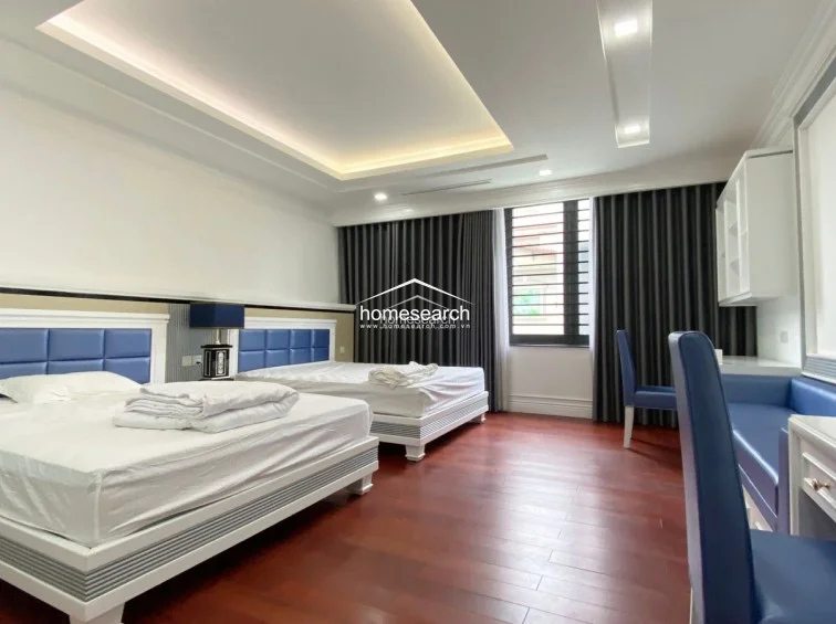 This bedroom is configured with two beds featuring blue upholstered headboards and a dedicated study area with a matching desk and chair. The room maintains a consistent aesthetic with dark wood flooring, neutral walls, and floor-to-ceiling dark curtains that frame the window.