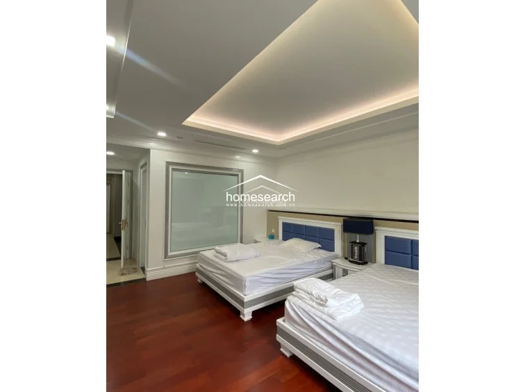 This bedroom layout mirrors the previous room, featuring two beds with blue upholstered headboards and a clean, minimalist design. The space is highlighted by a tray ceiling with warm ambient cove lighting and a large window that allows for soft, diffused natural light.
