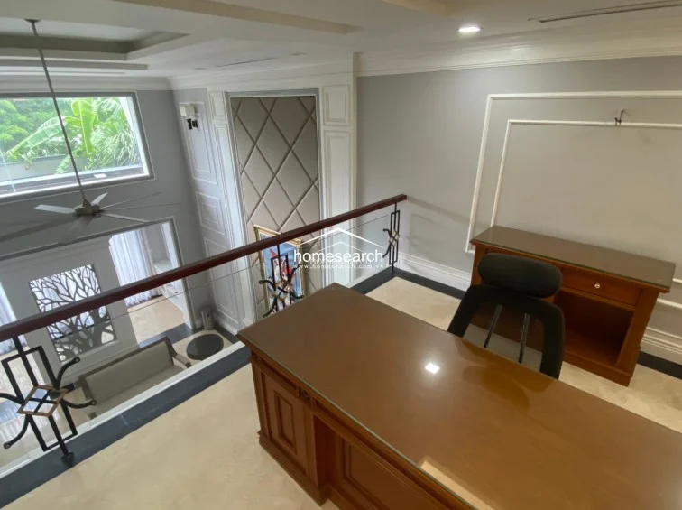 This mezzanine office space overlooks a living area, featuring a clean, modern layout with a glass-railed balcony and neutral grey walls with classic molding. The area is furnished with a professional wooden desk and chair, complemented by polished light-colored flooring and ample natural light from the large window.