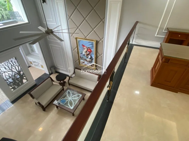 This high-ceilinged living area is viewed from a mezzanine level, highlighting an open-flow layout that connects the ground floor seating area to the upper landing. The space is decorated with a large statement painting, a modern ceiling fan, and a neutral color scheme, emphasizing a luxurious and well-maintained interior.