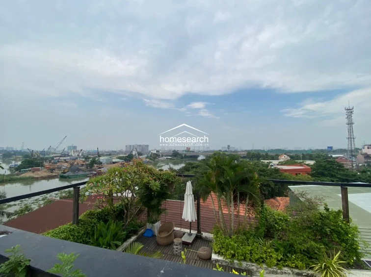 This rooftop terrace area is beautifully landscaped with integrated wooden planters filled with lush greenery and a small palm tree. It includes a dedicated seating nook with a parasol, providing a serene outdoor retreat with wide-reaching views of the river and urban skyline.