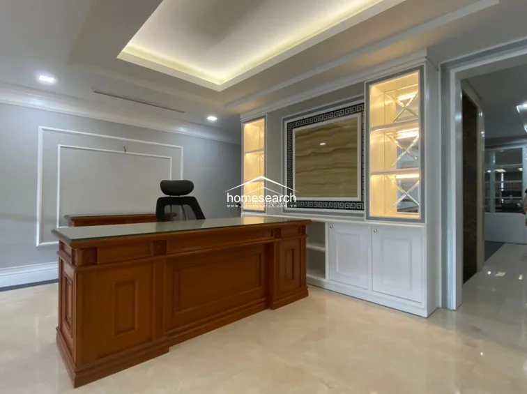 This professional office space features a high-quality wooden executive desk set against a sophisticated built-in wall unit with illuminated glass shelving and a decorative central panel. The room is finished with light-colored marble flooring, recessed ceiling lighting, and soft grey walls, creating a polished and formal atmosphere.
