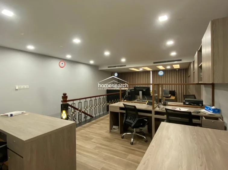 This open-plan office area features a modern, light-wood desk configuration with integrated partitions and ample storage cabinets. The space overlooks a staircase with a decorative metal railing and is illuminated by numerous recessed ceiling lights, maintaining a bright and productive environment.