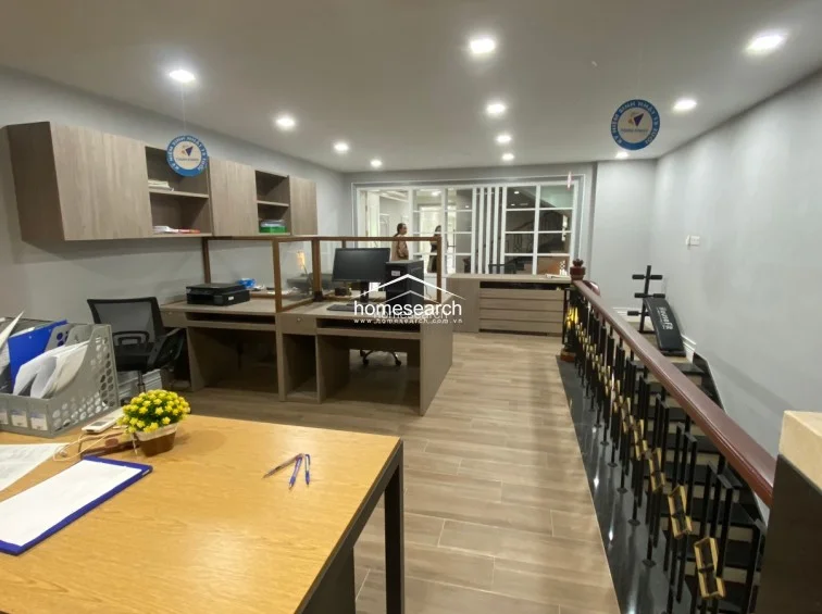 This functional office workspace is equipped with multiple desks, ergonomic chairs, and overhead storage units, all set against a backdrop of light-wood flooring. The area flows directly to an open staircase with a classic dark wood and metal balustrade, while a small fitness bench in the corner adds utility to the layout.
