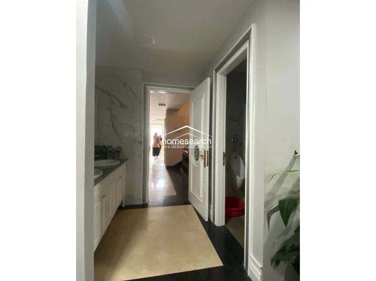 This hallway area features elegant marble flooring and connects to a bathroom equipped with a vanity and a separate water closet containing a urinal. The space is finished in a neutral color palette with white paneled doors and marble wall accents, reflecting a high level of maintenance.