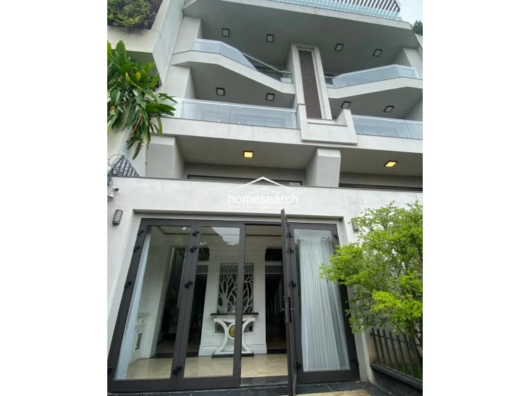 This exterior view showcases a multi-story building with contemporary architectural lines, featuring glass-railed balconies and large, dark-framed glass doors on the ground level. The property is well-maintained with integrated greenery and outdoor lighting, creating a sophisticated and welcoming facade.