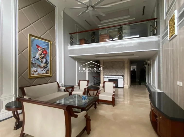 This grand, double-height living room is defined by a striking tufted accent wall featuring a large framed portrait and polished marble flooring. The layout includes a sophisticated seating arrangement, a sleek black media console, and a mezzanine level with a glass railing that enhances the sense of architectural openness.