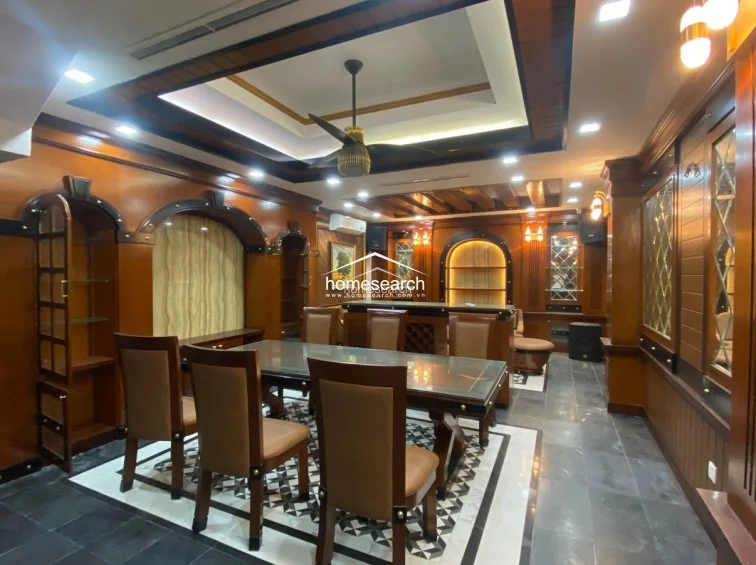 This formal dining or entertainment room is heavily accented with rich, polished wood paneling, built-in arched shelving, and a decorative coffered ceiling with a central fan. The floor features a striking combination of dark stone tiles and a central geometric patterned inlay, creating a luxurious and traditional atmosphere.