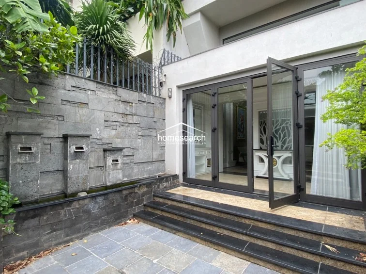 This outdoor terrace area features a sophisticated water feature built into a textured stone wall, creating a serene and private atmosphere. The space connects seamlessly to the interior living area through large glass folding doors, with steps leading up to the main floor, all finished in high-quality stone and tile.