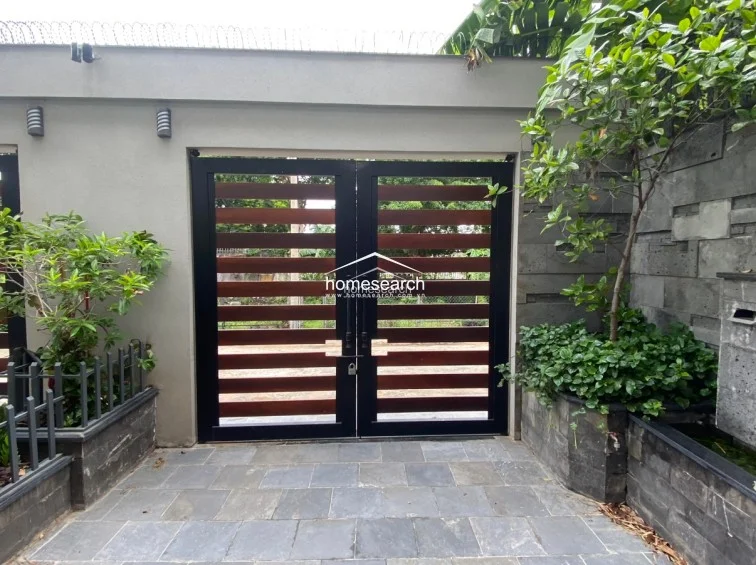 This private entrance area features a secure, modern gate made of horizontal wood slats framed in black metal, set against a concrete wall. The space is enhanced by stone-tiled flooring and lush landscaping, including a small water feature and potted plants, providing a clean and well-kept arrival experience.