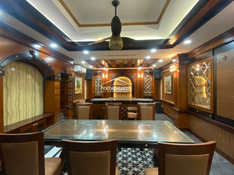 This dining or lounge space is defined by extensive woodwork, including wall-to-wall paneling, built-in display cabinets with mirrored accents, and an elaborate recessed ceiling. The room is equipped with a wall-mounted AC unit, recessed lighting, and a large glass-topped table, reflecting a high level of maintenance and a formal, classic interior design.