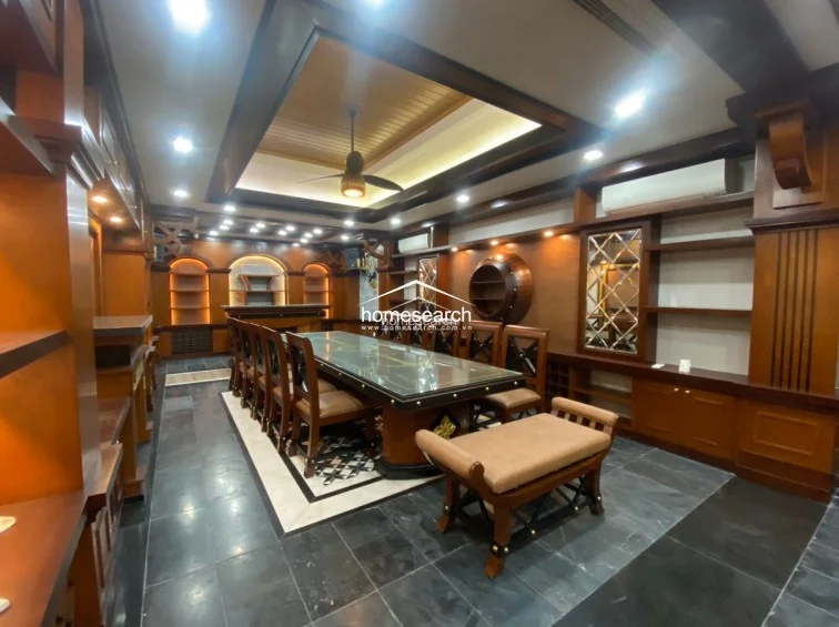 This formal dining or meeting room features a large, long table with matching chairs, set against a backdrop of extensive, high-quality custom wood cabinetry and shelving. The space is defined by a sophisticated coffered ceiling with integrated lighting, a ceiling fan, and dark stone flooring with a decorative geometric inlay.