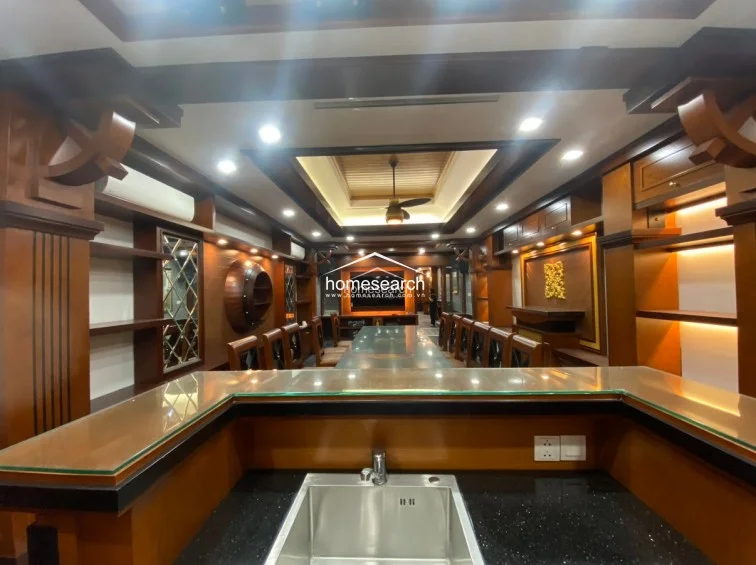 This expansive room features a sophisticated, dark wood-paneled design with a long dining or conference table and integrated shelving units. The ceiling includes intricate recessed lighting and a central fan, while the space flows into a bar area with a black stone sink, creating a luxurious and formal atmosphere.