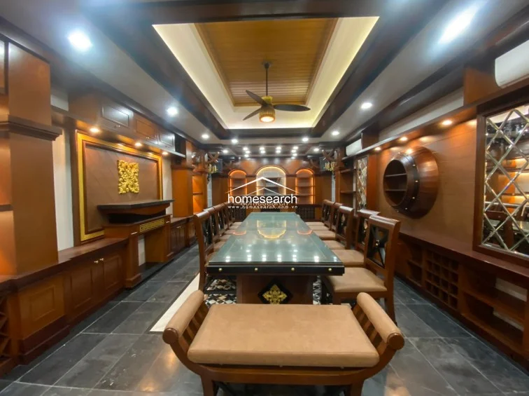 This expansive formal dining room is dominated by a long, glass-topped table surrounded by numerous wooden chairs, suggesting a space designed for large gatherings. The room is heavily detailed with rich wood cabinetry, decorative gold accents, a coffered ceiling with a central fan, and integrated ambient lighting that highlights the luxurious, traditional aesthetic.