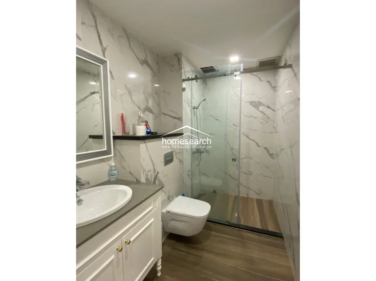 This modern bathroom features elegant white marble-veined wall tiles and a sleek glass-enclosed shower area with a rainfall showerhead. The vanity includes a white basin set into a grey stone countertop, complemented by a wall-mounted toilet and warm recessed lighting.
