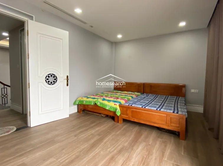 This bedroom features a minimalist design with light grey walls, wood-look tile flooring, and two wooden bed frames placed side-by-side. The room includes a distinctive white door with a decorative circular inlay and is serviced by recessed ceiling lights and a concealed air conditioning vent, presenting a clean and well-maintained living space.