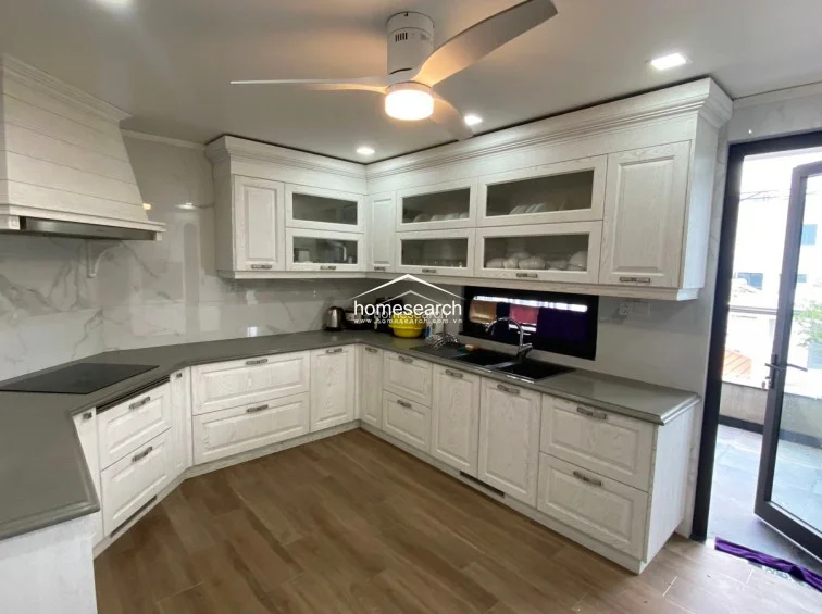 This U-shaped kitchen features light-toned, textured cabinetry, dark grey countertops, and a modern induction cooktop with a matching range hood. The space is finished with wood-look tile flooring and a marble-patterned backsplash, with a glass door providing direct access to an outdoor area for added convenience.