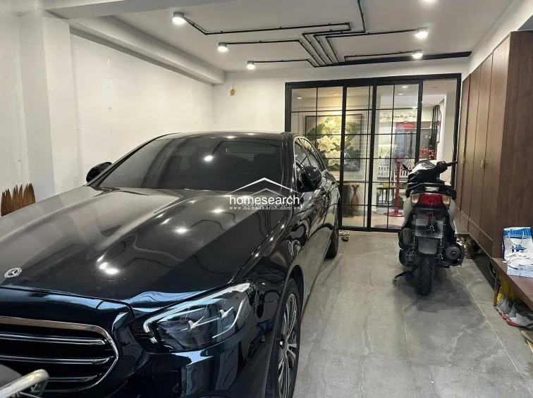 This spacious garage area features large-format grey floor tiles and a minimalist industrial ceiling design with exposed black conduit lighting. A full-height wooden storage cabinet lines the right wall, while a sleek black-framed glass partition provides a visual connection to the interior living space.