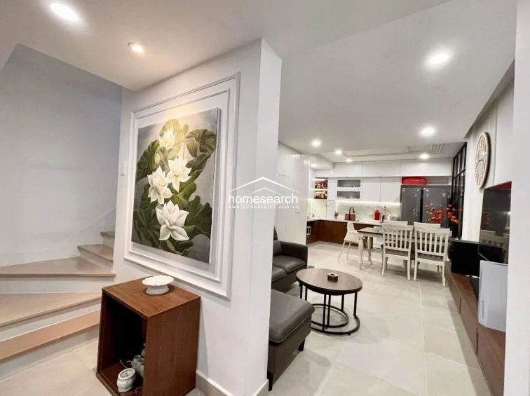 This open-plan living and dining area is anchored by a striking floral wall art piece and a wooden console, leading directly into a modern kitchen with white cabinetry. The space features light-colored tile flooring, recessed ceiling lights, and a neutral color palette that creates a bright, cohesive flow.