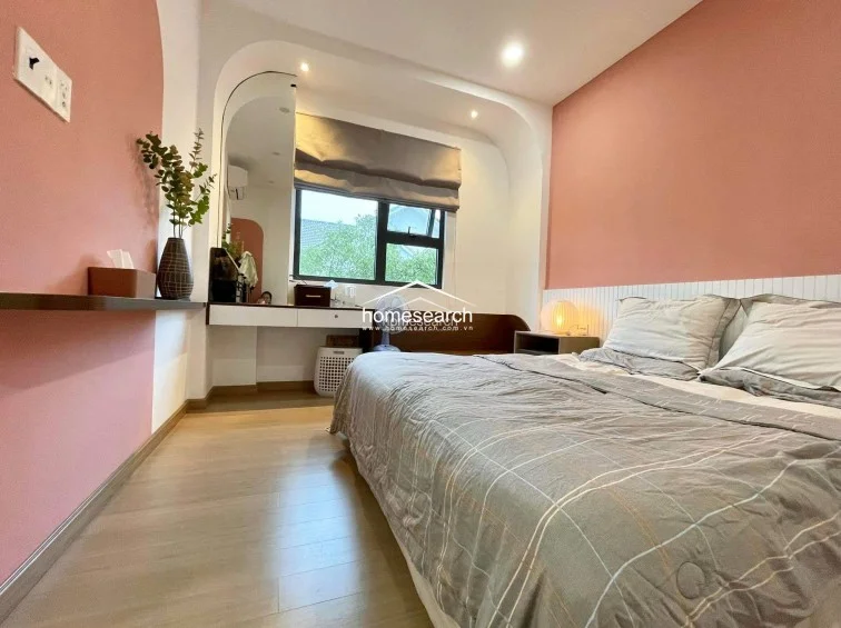This bedroom showcases a creative design with soft pink walls and an elegant arched architectural detail framing the vanity area. The room is equipped with light wood flooring, a built-in floating desk, and a window with a grey Roman shade that offers a pleasant view of the outdoors.
