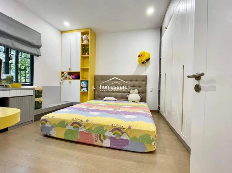 This vibrant bedroom is designed with a playful aesthetic, featuring a yellow-accented storage unit and a colorful rainbow-patterned bedspread. It includes extensive white built-in wardrobes for ample storage, light wood flooring, and a large window that invites natural light into the room.
