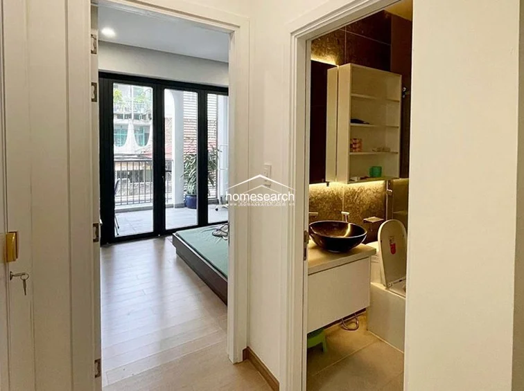 This image shows a transitional hallway connecting a bedroom to a modern bathroom. The bathroom features a stylish dark-toned vessel sink, white vanity, and warm ambient lighting, while the bedroom beyond boasts light wood flooring and large glass doors leading to a balcony.