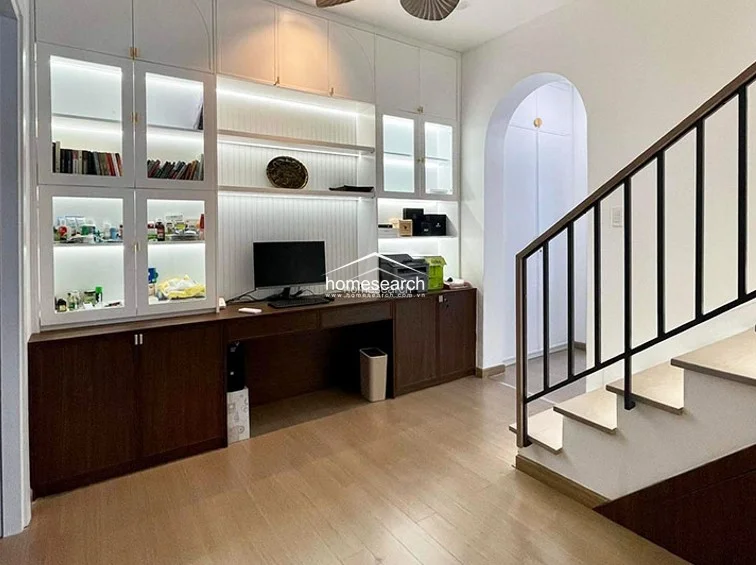 This functional home office or study area features extensive custom floor-to-ceiling cabinetry with integrated LED lighting and a built-in desk. The space is anchored by a staircase with a modern black metal railing and a sophisticated arched entryway, all finished with light wood flooring and neutral walls.