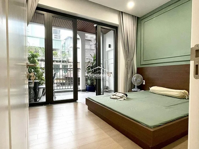 This bedroom offers a serene atmosphere with sage green wall paneling, a low-profile dark wood bed frame, and light wood flooring. Large black-framed glass doors provide excellent natural light and direct access to a private balcony, which is furnished with potted plants and outdoor seating.