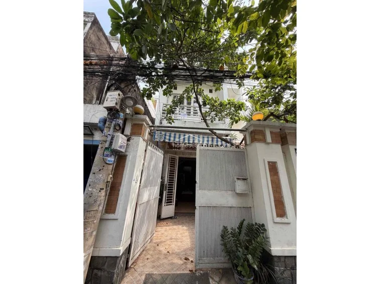 This exterior view shows the front facade of a multi-story townhouse, featuring a gated entrance with a small courtyard area. The property includes a balcony on the upper level and is shaded by a mature tree, though overhead utility lines are prominent.