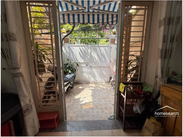 This perspective from inside looking out shows the transition between the interior living space and the gated courtyard, which serves as a secure parking area for motorbikes. The space is protected by a retractable striped awning and features tiled flooring and metal security grilles.