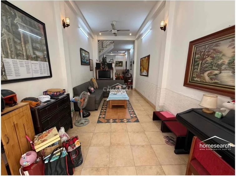 The living room features a long, narrow layout with light-colored tile flooring and a central decorative tile inlay. The space is furnished with a large grey sectional sofa and connects directly to the rear dining and kitchen area, with a staircase visible in the background.