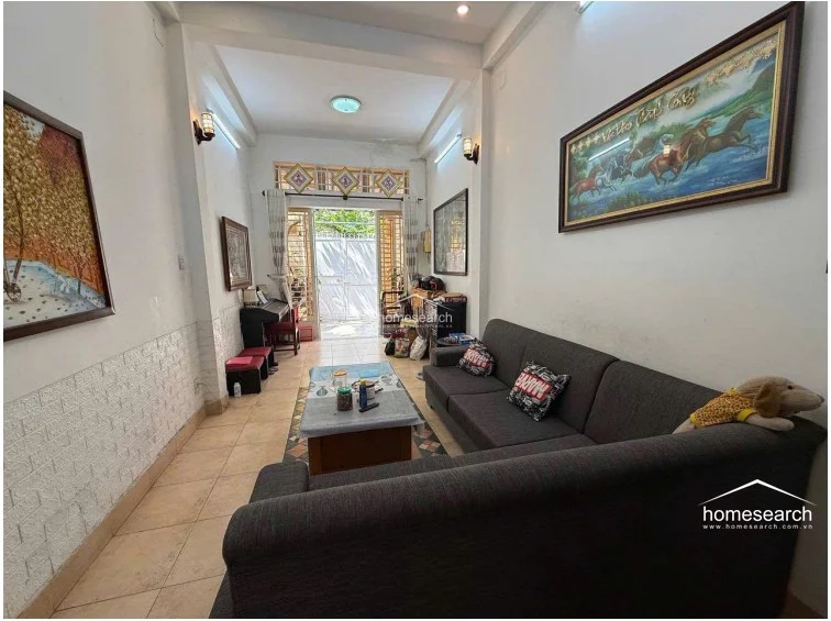 This reverse angle of the living room highlights the connection to the front courtyard through a glass-paned door, allowing for natural light to filter into the space. The walls are painted a neutral white, accented by large framed artwork and wall-mounted light fixtures.