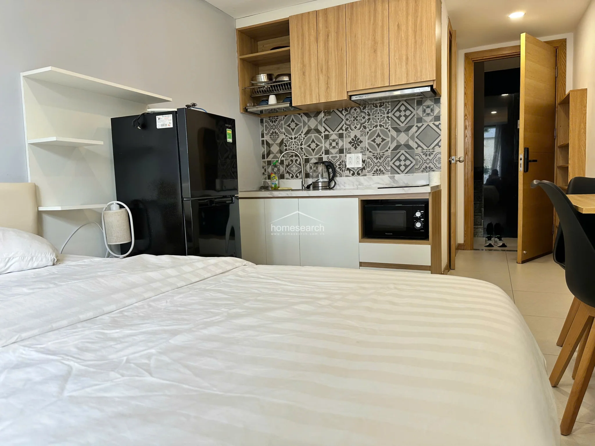 The compact kitchenette is efficiently integrated into the bedroom space, featuring light wood cabinetry, a microwave, and a black mini-fridge. The area is accented by a stylish patterned tile backsplash and a clean, modern aesthetic that maintains a cohesive flow with the rest of the unit.