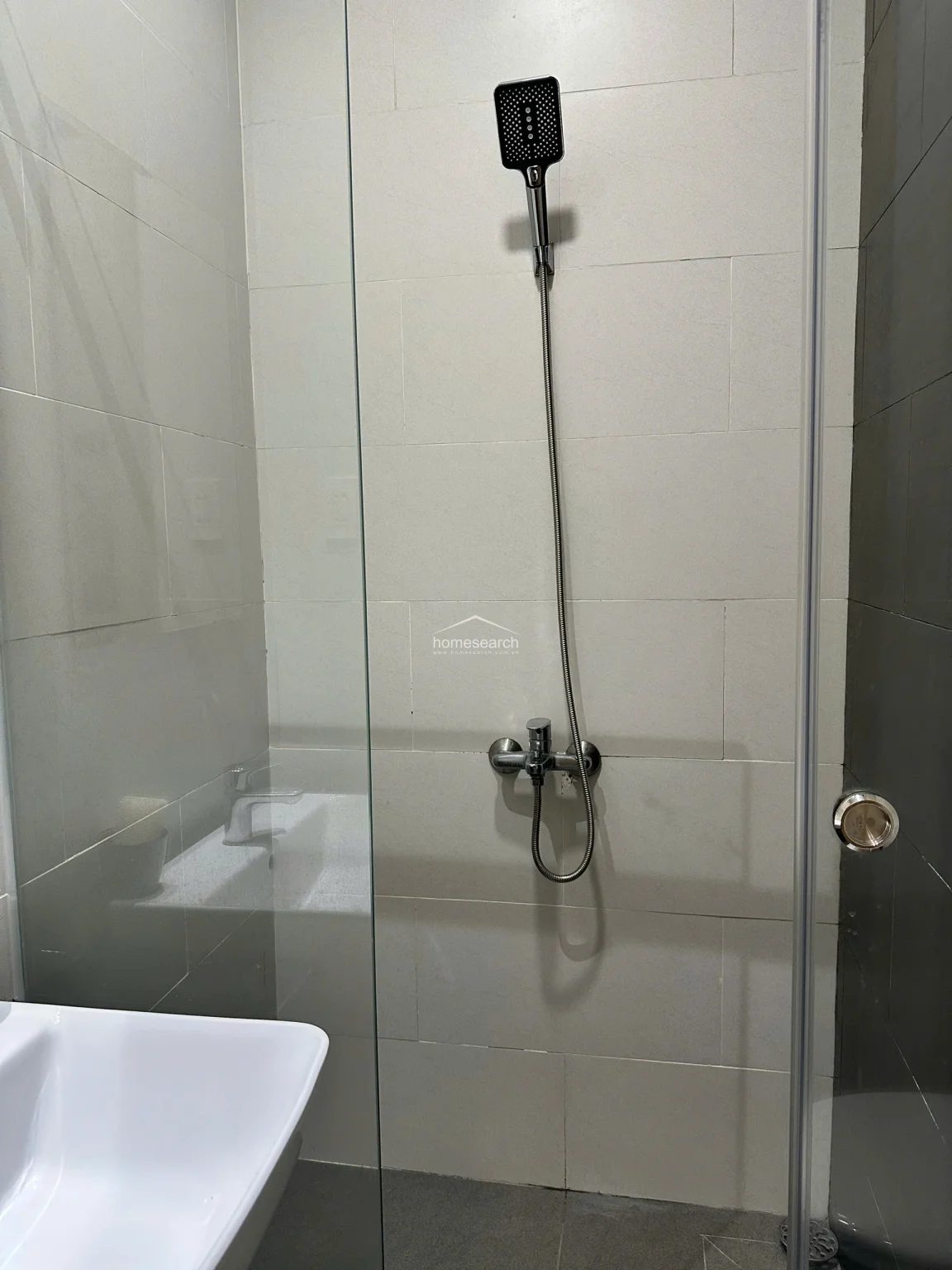 The bathroom is finished with neutral, large-format wall tiles and features a contemporary glass-enclosed shower stall. It is equipped with a modern square showerhead and a clean white vanity, reflecting a high level of maintenance and a functional, minimalist design.