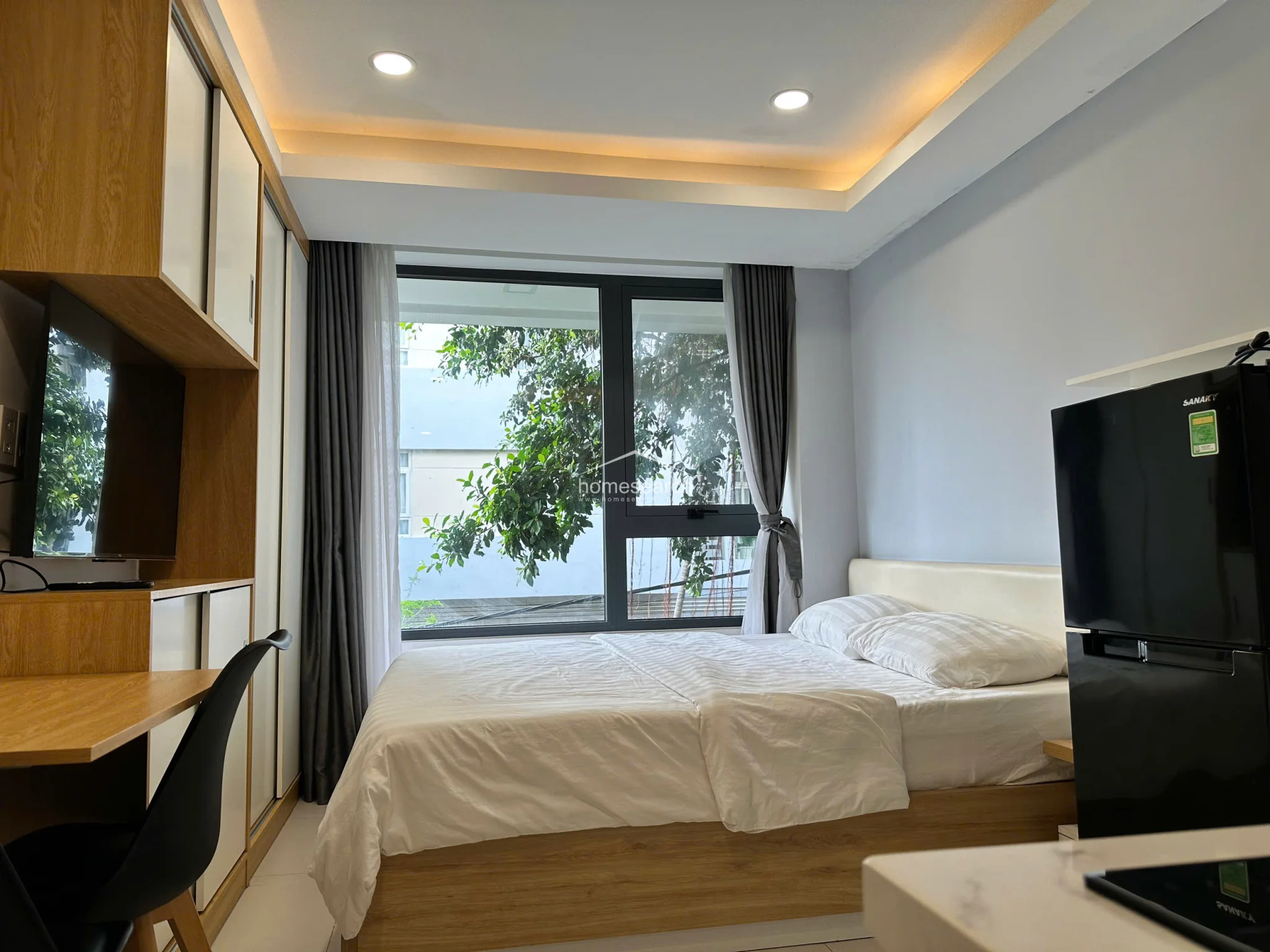 This wide-angle view highlights the studio's efficient layout, showcasing the seamless transition between the sleeping area, the workspace, and the kitchenette. The room is well-maintained with light-colored flooring, soft ambient cove lighting, and a modern, minimalist color scheme that maximizes the feeling of space.