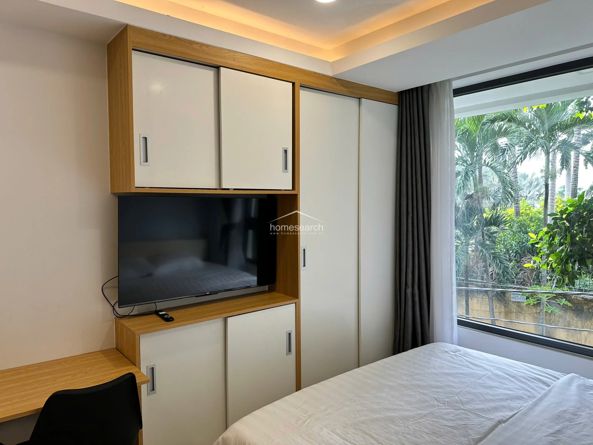 This studio bedroom features a functional, space-saving design with a built-in wooden wardrobe and integrated TV console. A large window provides ample natural light and a pleasant view of surrounding greenery, complemented by modern recessed ceiling lighting and neutral wall finishes.