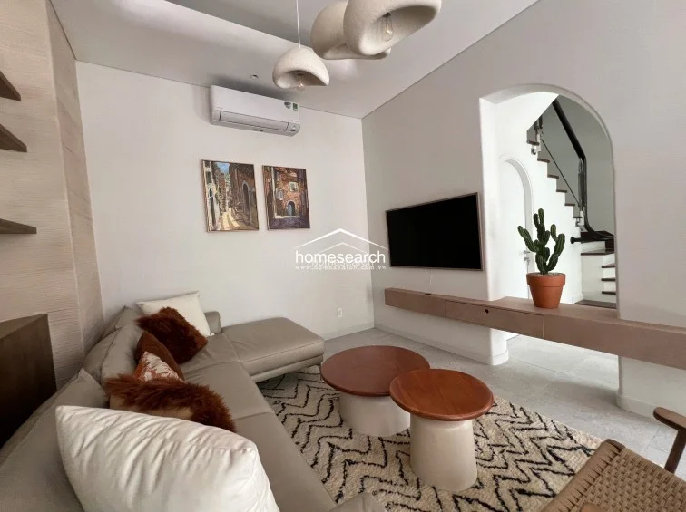This living area features a modern, minimalist aesthetic with a neutral color palette and soft, rounded architectural elements like the arched entryway. It is equipped with a wall-mounted AC unit, a large flat-screen TV, and a floating media console, all set against clean white walls and light-toned flooring. The space flows directly into the staircase area, creating an open and airy feel enhanced by contemporary pendant lighting.