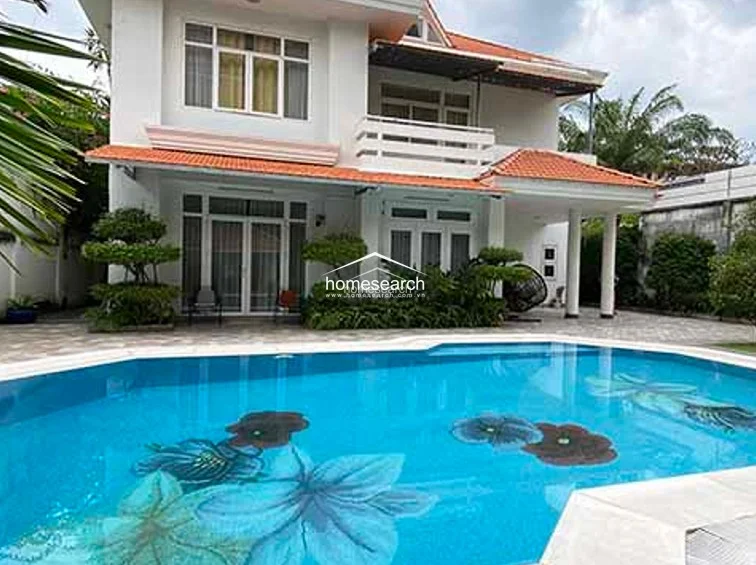 This two-story villa features a classic architectural style with a vibrant orange-tiled roof, white exterior walls, and a prominent second-floor balcony. The property is centered around a large, custom-tiled swimming pool adorned with floral mosaic patterns, set against a backdrop of lush, manicured greenery and mature trees.