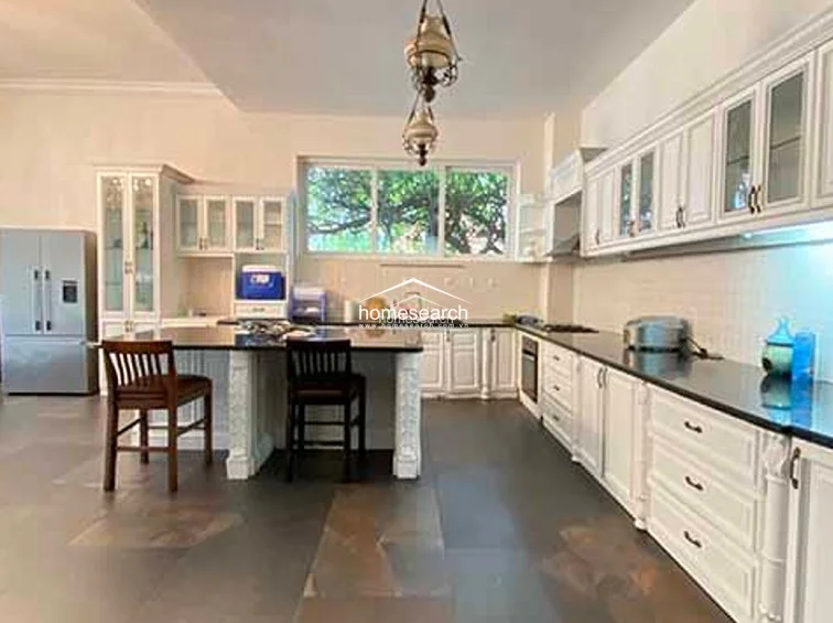 This spacious kitchen and dining area features a large central island with bar seating, illuminated by a decorative, multi-tiered pendant light fixture. The space is finished with high-quality white cabinetry, integrated stainless steel appliances, and dark, large-format floor tiles that provide a sophisticated contrast to the light walls.