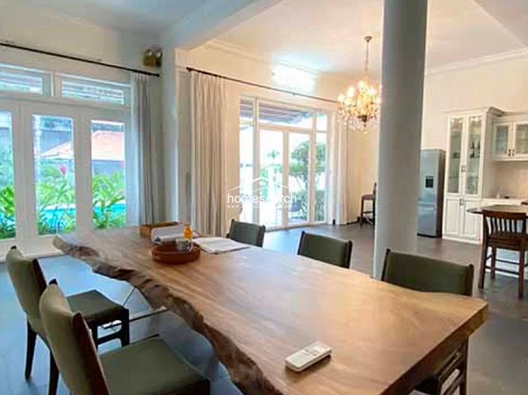 The dining room is anchored by a stunning, large-slab live-edge wooden table, complemented by modern, dark-upholstered chairs. The room is flooded with natural light from expansive floor-to-ceiling windows and doors, which offer a clear view of the exterior garden and pool area, while a grand central column adds a structural architectural detail to the open-plan space.
