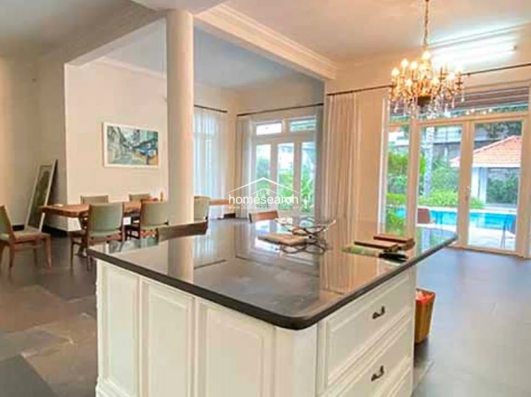 This spacious interior room serves as a formal dining or multi-purpose area, featuring a large kitchen island with a dark stone countertop and a decorative crystal chandelier. The space flows seamlessly to the outdoors through floor-to-ceiling glass doors, which provide abundant natural light and direct views of the swimming pool area.