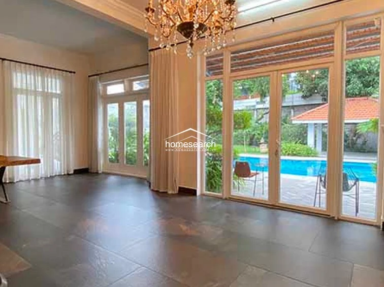 This interior space features large-format dark tile flooring and floor-to-ceiling glass doors that open directly onto the pool deck, emphasizing an indoor-outdoor living flow. The room is illuminated by a grand crystal chandelier and benefits from bright natural light, with neutral wall finishes and elegant floor-length curtains.