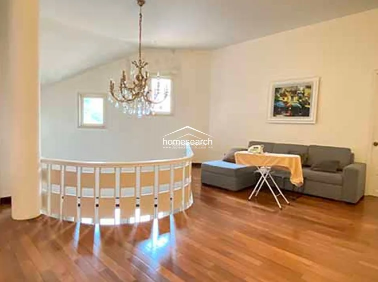 This upper-level landing serves as a cozy secondary living area, featuring warm hardwood flooring and a classic crystal chandelier. The space is defined by a curved, white-paneled railing that overlooks the main floor, creating an open and airy architectural flow.