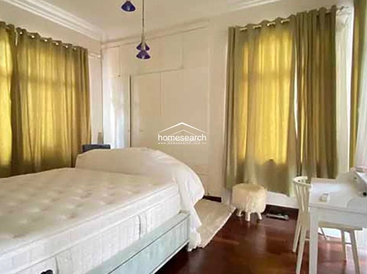 This bedroom features a clean, minimalist aesthetic with neutral wall tones and warm hardwood flooring. It is equipped with floor-to-ceiling built-in wardrobes for ample storage and is accented by soft, floor-length gold curtains that add a touch of elegance.