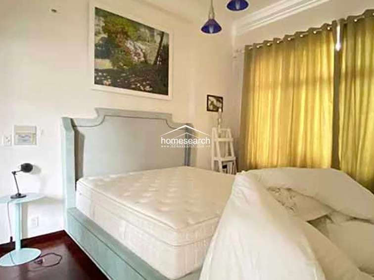 This bedroom centers on a large, upholstered light-grey bed frame that provides a soft, modern focal point against the neutral walls. The room maintains a consistent design language with the rest of the home, featuring hardwood floors, gold drapery, and a decorative art piece above the headboard.