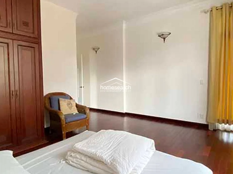 This bedroom features warm hardwood flooring, neutral wall tones, and a large built-in wooden wardrobe for ample storage. A classic armchair sits in the corner, while wall-mounted sconces and a window draped in soft yellow curtains create a cozy, well-maintained atmosphere.