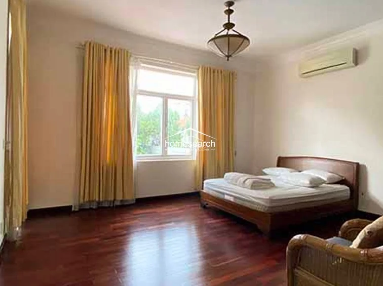 This bedroom is defined by its minimalist aesthetic, featuring a large bed, hardwood flooring, and floor-to-ceiling yellow curtains that frame the window. A decorative pendant light hangs from the ceiling, and a wall-mounted air conditioning unit ensures climate control in this clean, well-kept space.