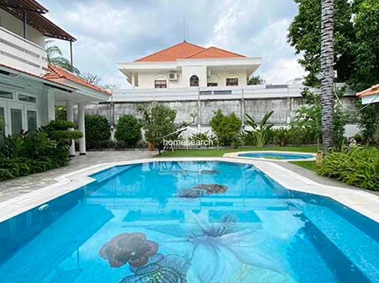 This outdoor space features a private swimming pool with a unique, decorative floral mosaic inlay at the bottom. The surrounding area includes a paved terrace, lush greenery, and a view of a neighboring multi-story residence under a cloudy sky.