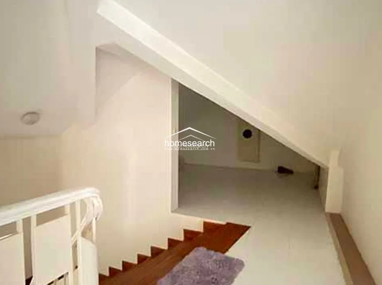 This image shows an upper-level landing or mezzanine area with a white wooden staircase railing and a small, cozy attic nook. The space is finished with light-colored flooring and a small decorative rug, highlighting the architectural interest of the sloped roof design.